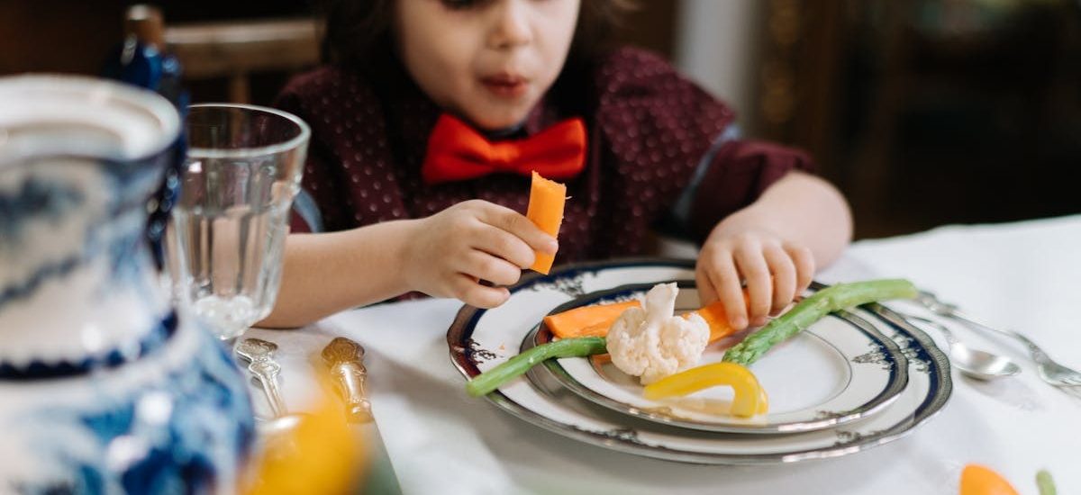 A child in a red bow tie enjoys a healthy meal with vegetables indoors.