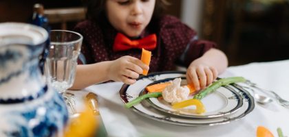 A child in a red bow tie enjoys a healthy meal with vegetables indoors.