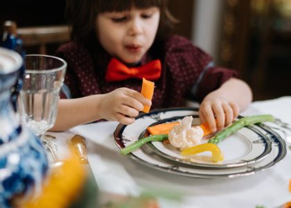 A child in a red bow tie enjoys a healthy meal with vegetables indoors.